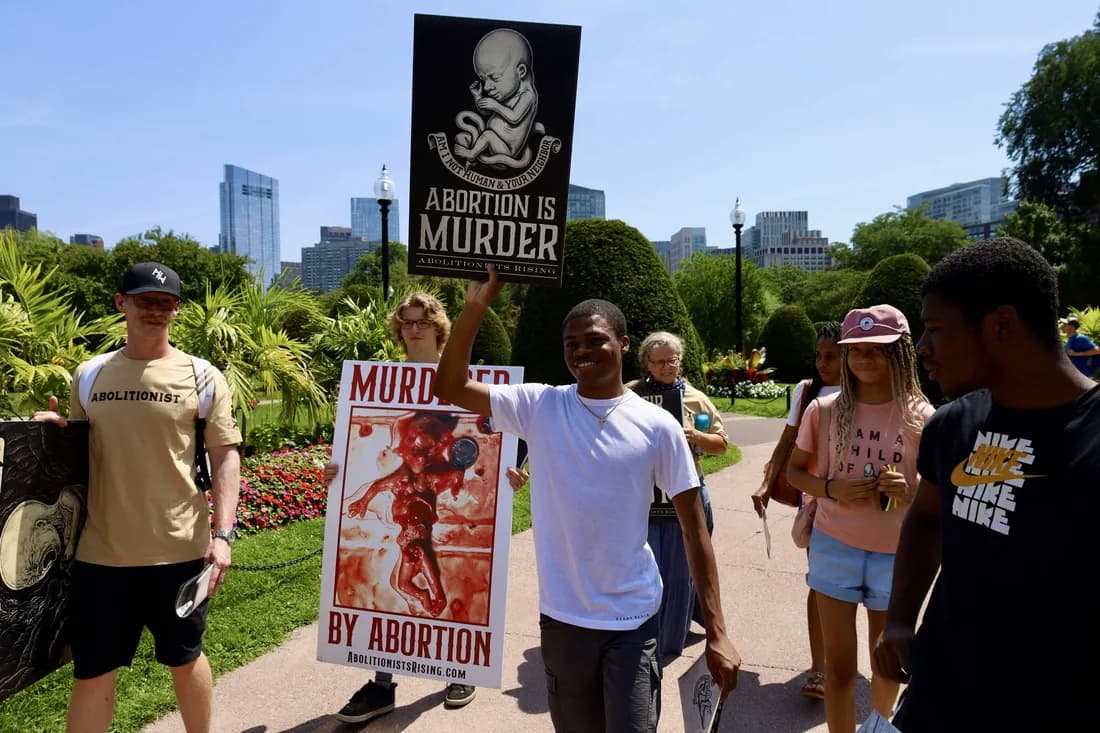 A young man raising an "Abortion is murder" sign on a park path as a group walks alongside him.